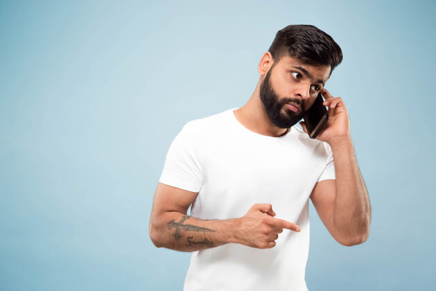 工作蓝色背景上穿着白衬衫的印度教年轻人的半身特写肖像人类情感 面部表情 广告概念负面空间打电话 录音留言衬衫手势年轻