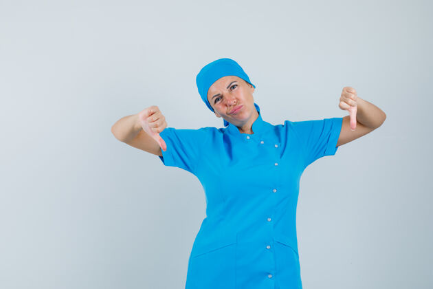 疾病女医生穿着蓝色制服 双拇指朝下 看上去很失望 正视图成人护士失望