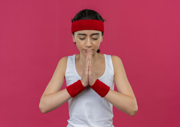 表情身穿运动服的年轻健身女 头上戴着头巾 双臂抱在一起 带着希望的表情 闭着眼睛站在粉色的墙上祈祷运动装姿势手臂