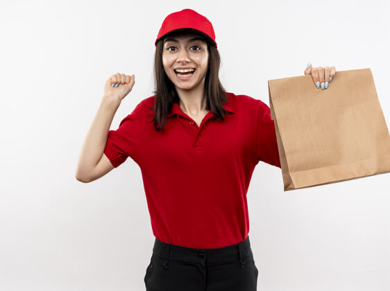 举行年轻的送货员身穿红色制服 戴着帽子 手里拿着纸包 握紧拳头 站在白色的背景上 开心而兴奋年轻帽子握紧