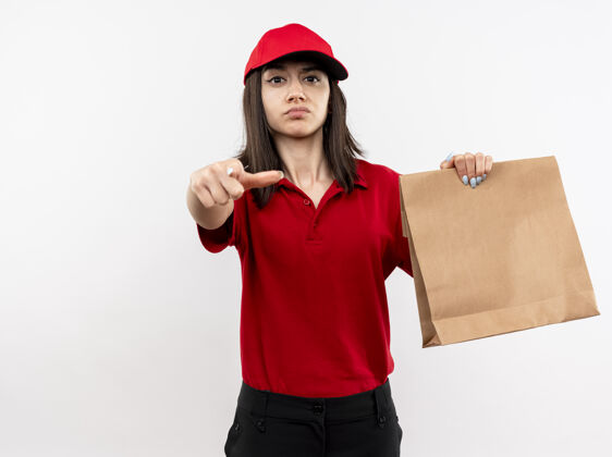 目录身穿红色制服 戴着帽子的年轻送货女孩拿着纸包 用食指指着镜头 严肃的脸站在白色的背景上手指拿着脸