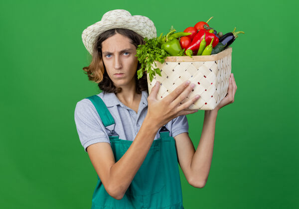 花园年轻的园丁穿着连体衣戴着帽子拿着装满新鲜蔬菜的箱子充分帽子严重