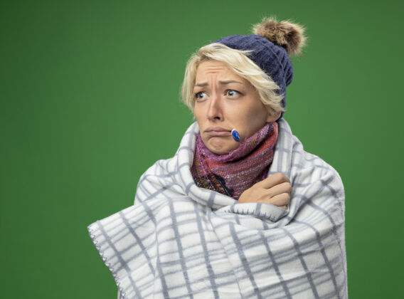 温暖生病的不健康的妇女 短头发 戴着暖和的围巾和帽子 嘴里拿着温度计站在绿色背景上 感觉不舒服围巾帽子站