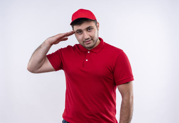 帽子身穿红色制服 头戴鸭舌帽的年轻送货员站在白墙上自信地微笑致意公民制服姿势