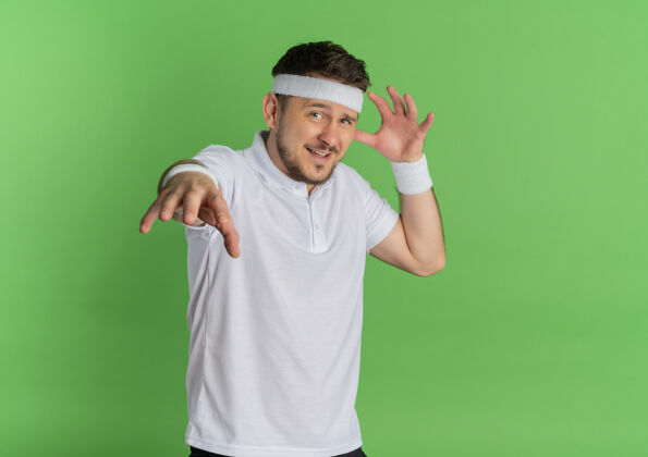 男人身穿白衬衫 头戴头巾的年轻健身男子站在绿色的墙壁上 快乐而积极地伸出双手望着前方头带站积极