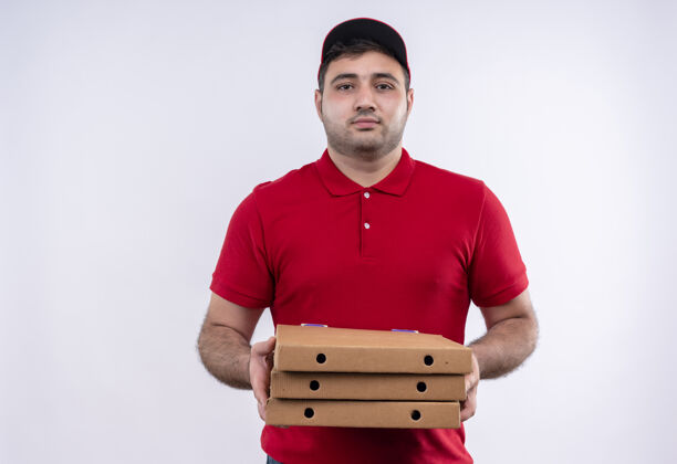 姿势年轻的送货员身穿红色制服 戴着帽子 手里拿着披萨盒 站在白色的墙上 自信地看着镜头拿着人自信