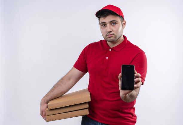 表情年轻的送货员身穿红色制服 头戴鸭舌帽 拿着披萨盒 站在白色的墙上 表情自信地展示着智能手机站着表演男人