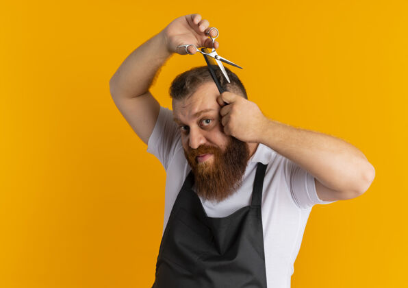 头发专业的胡须理发师站在橙色的墙上 围着围裙用剪刀剪头发胡子男人家伙
