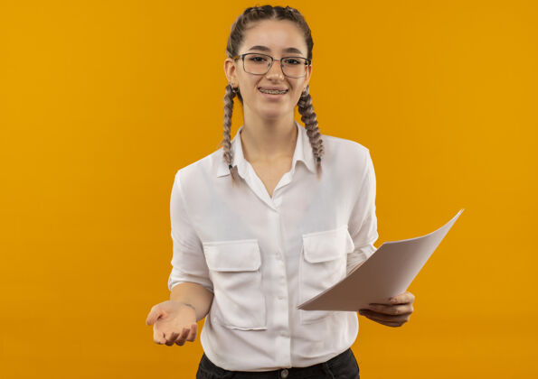 拿着戴着眼镜 梳着辫子 穿着白衬衫的年轻女学生 手里拿着空白的书页 面带微笑地站在橙色的墙上 望着前面欢呼手势姿势