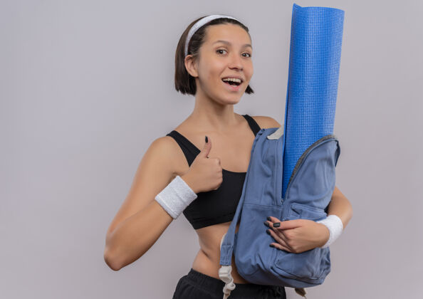 微笑穿着运动服的年轻健身女士背着背包 脸上挂着瑜伽垫 微笑着竖起大拇指站在白色的墙上年轻运动健康
