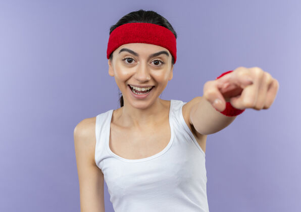 头带身穿运动服的年轻健身女 头上戴着头巾 微笑着 用食指指着站在紫色墙上的摄像机目录站立欢呼