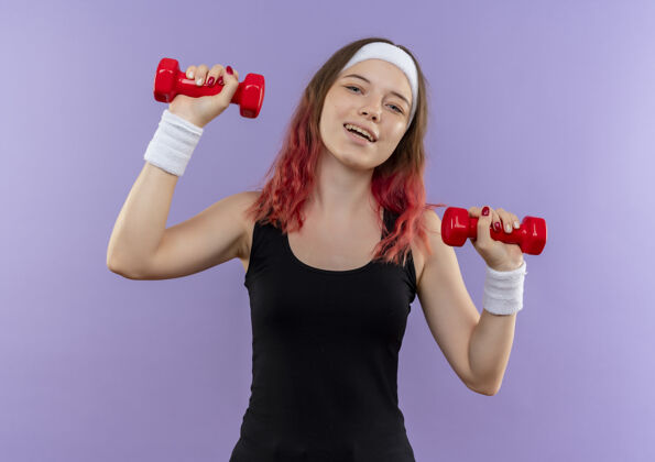 运动员穿着运动服的年轻健身女士正在用哑铃做练习站在紫色的墙上开心地笑运动市民姿势