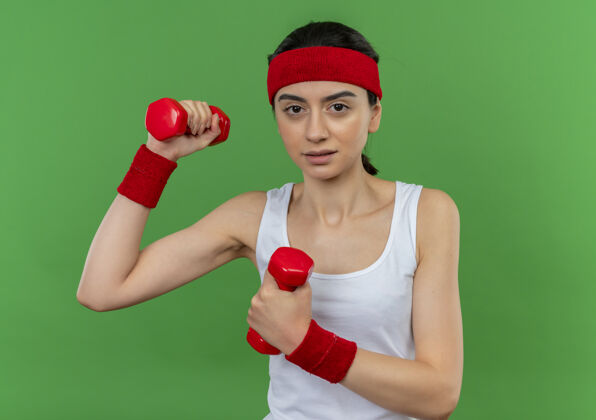 看身穿运动服的年轻健身女士 头戴钢带 举着两个哑铃 自信地举手练习 严肃的脸站在绿色的墙上哑铃健身运动