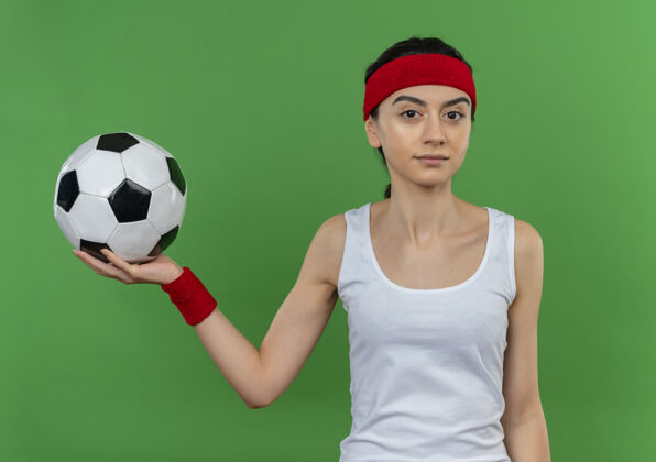 站着身穿运动服 头箍 手持足球 表情自信的年轻健身女士站在绿色的墙上运动员表情健身