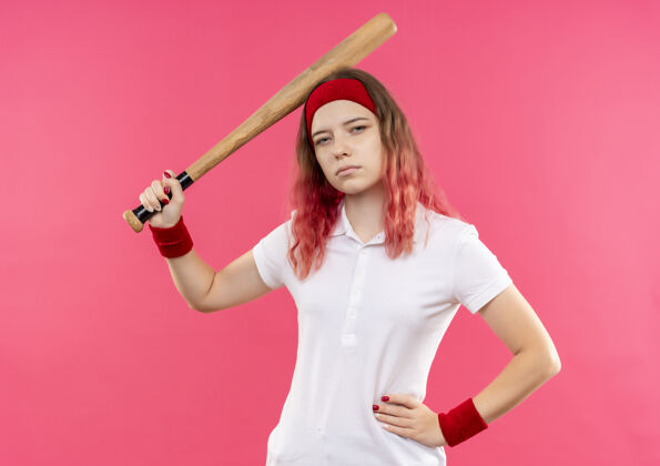 运动装戴着头巾打棒球的年轻运动型女子手持球棒 站在粉红色的墙上 看上去很自信人抱运动员
