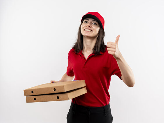 拿着身穿红色制服 头戴鸭舌帽 拿着披萨盒的年轻送货员看着镜头 满脸笑容 站在白色背景上竖起大拇指帽子拇指制服