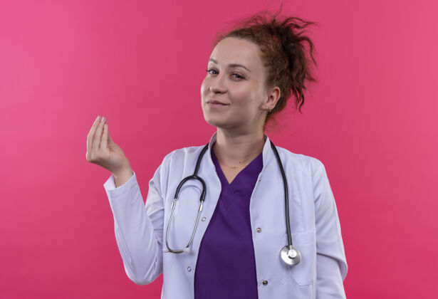 听诊器年轻的女医生穿着白大褂用听诊器搓手指赚钱的手势用手看着自信的要钱站在粉红色的墙上看女人做
