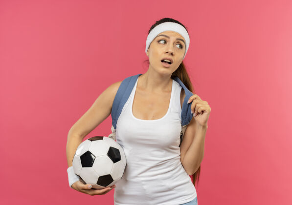 头带身穿运动服的年轻健身女士 头上戴着头带 脖子上挂着金牌 背着背包 手里拿着足球 站在粉色的墙上 困惑地看着一边运动装旁边人