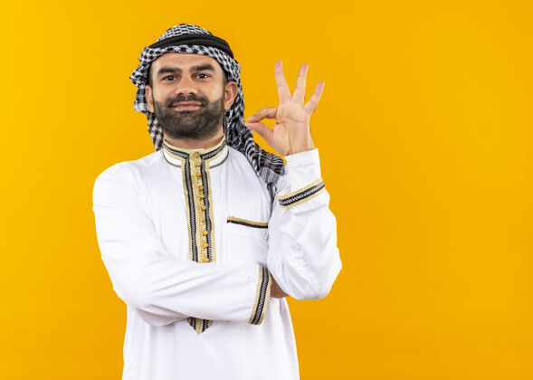 站身着传统服装的阿拉伯商人微笑自信地站在橙色的墙上做着“ok”的手势微笑阿拉伯语穿