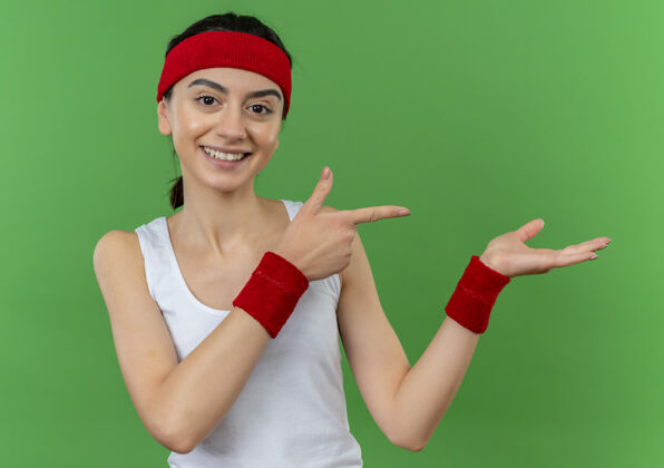 姿势身穿运动服的年轻健身女士 头戴花环 微笑着 自信地用食指指着侧面 手臂站在绿色的墙上人手臂手