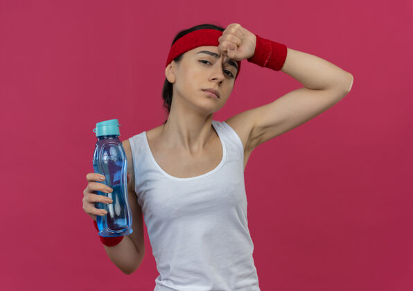 水穿着运动服的年轻健身女 头上戴着头巾 手里拿着一瓶水 站在粉红色的墙上 看上去又累又累健身人疲倦