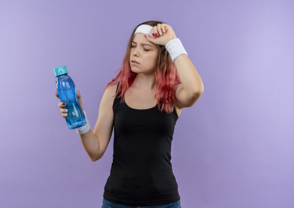 瓶子穿着运动服的年轻健身女士站在紫色的墙上 手里拿着一瓶水 看起来很累看女性累