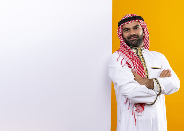 空白身着传统服装的阿拉伯商人站在空白的墙板旁 脸上带着自信的笑容 站在橙色的墙上站微笑广告牌