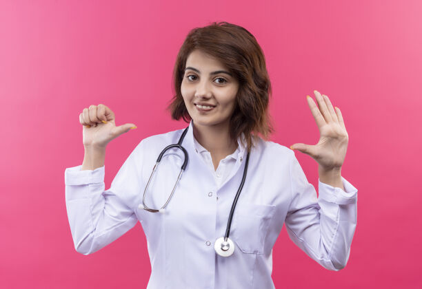 女人身穿白大褂 手持听诊器的年轻女医生站在粉红色的墙上 手指朝上 手指朝上 微笑着医生外套手指