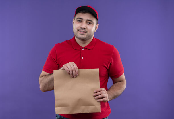 微笑年轻的送货员身穿红色制服 头戴鸭舌帽 面带微笑 站在紫色的墙上年轻人包装递送