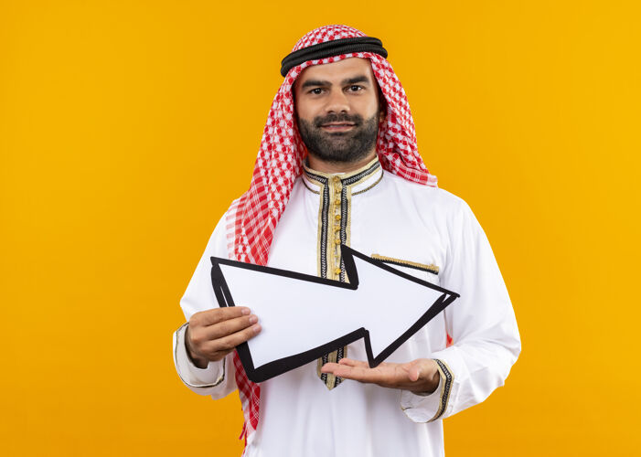 脸身着传统服饰的阿拉伯商人手持指向右边的大箭头 脸上带着微笑站在橙色的墙上传统指箭