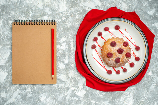 浆果蛋糕俯视图白色椭圆形盘子上的浆果蛋糕红色披肩红色铅笔笔记本上的灰色表面椭圆形视图披肩