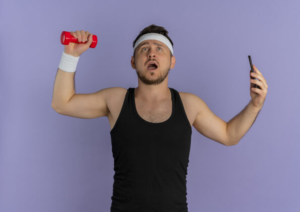 小伙子戴着头巾的年轻健身男子用智能手机自拍 手里拿着哑铃站在紫色的墙上健康哑铃健身