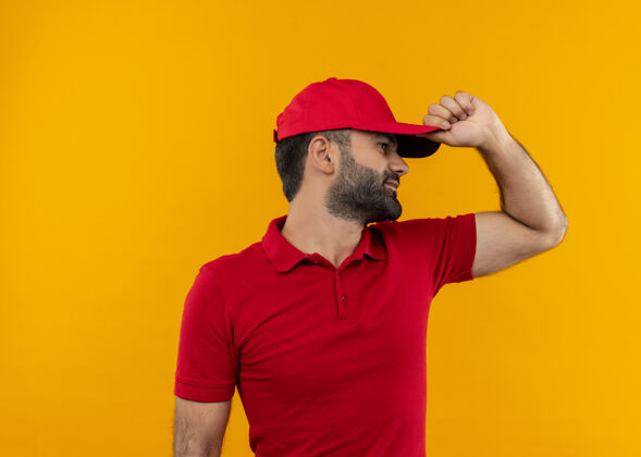 送货一个留着胡须的送货员 穿着红色制服 戴着帽子 手放在帽子上 向旁边看 看有人站在橙色的墙上靠边站制服