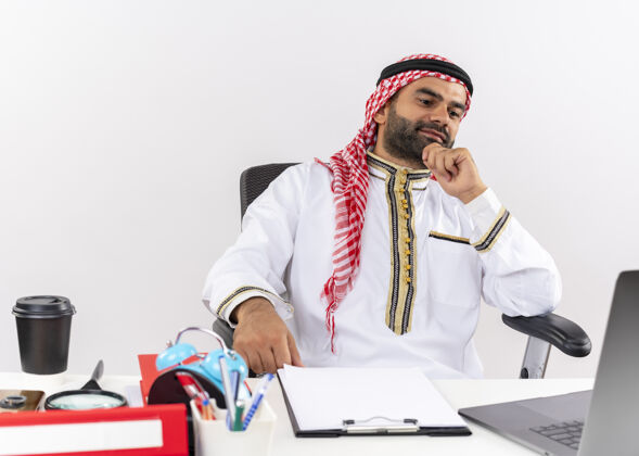 传统身着传统服饰的阿拉伯商人坐在桌旁 用笔记本电脑思考 用沉思的表情在办公室工作桌子坐商务人士