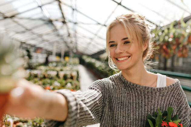 花可爱的绿眼睛微笑的女人的肖像在温室里摆姿势并且拿着植物自然金发毛衣