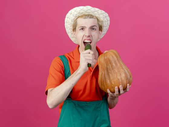 男人年轻的园丁 穿着连体衣 戴着帽子 手里拿着南瓜和黄瓜抱着穿黄瓜