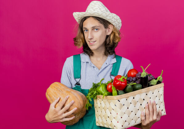 微笑年轻的园丁穿着连体衣戴着帽子拿着装满新鲜蔬菜的箱子男人帽子年轻