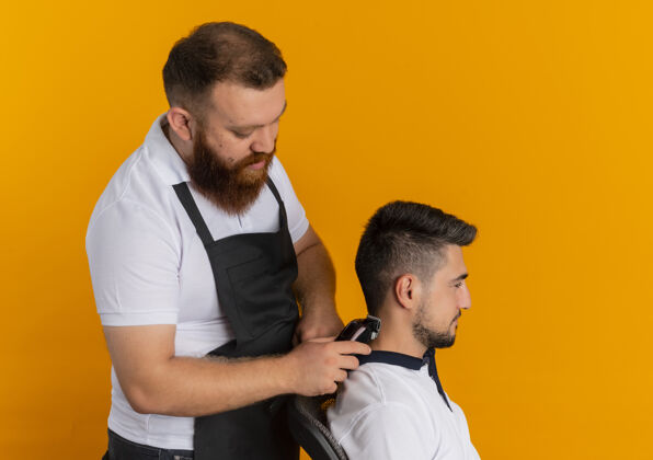 男人专业的胡须理发师围着围裙用剃须刀机给站在橘色墙上的年轻人理发围裙机器站着