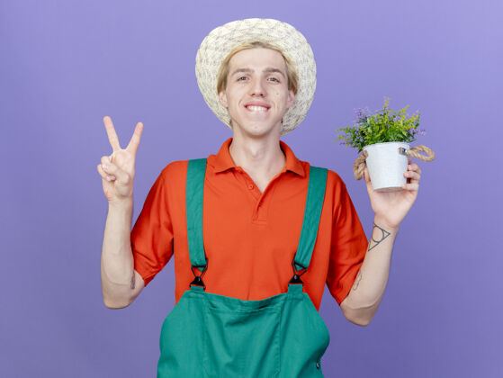站着年轻的园丁穿着连体衣 戴着帽子 手里拿着盆栽植物微笑着Vsign抱着锅