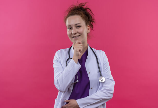 外套年轻的女医生穿着白大褂 手放在下巴上 微笑着站在粉红色的墙上下巴微笑医生