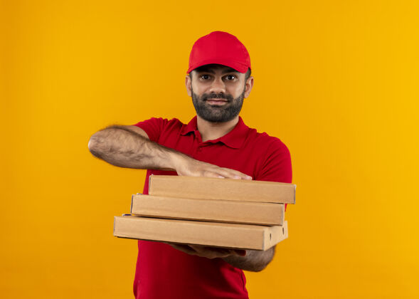 叠着身穿红色制服 头戴鸭舌帽 满脸胡须的送货员站在橙色的墙上 手里拿着一叠披萨盒 面带友好的微笑盒子提议男人