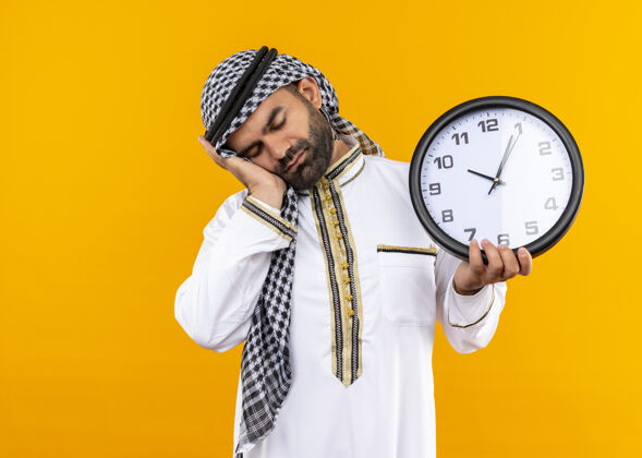 拿着穿着传统服装的阿拉伯商人拿着挂钟 头靠在手掌上 看起来很累 站在橙色的墙上想睡觉累了穿传统