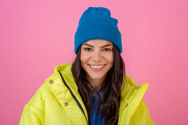 秋天一幅迷人微笑的时尚女人的画像 穿着黄色的彩色冬季羽绒服 戴着蓝色的针织帽子 穿着暖和的衣服 在粉色的墙上摆姿势 时尚潮流温暖运动户外