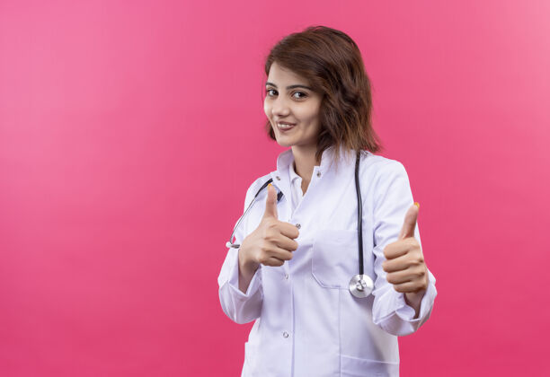 听诊器身穿白大褂 手持听诊器的年轻女医生微笑着 双手竖起大拇指站在粉红色的墙上拇指欢呼两者