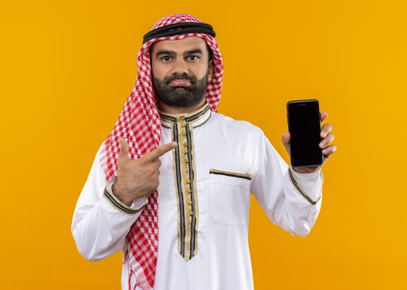 站身着传统服装的阿拉伯商人站在橙色的墙上 用手指指着智能手机 看上去很困惑指困惑手指