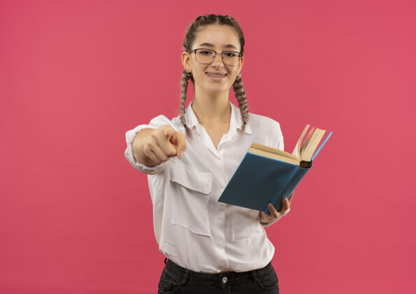 握着戴着眼镜 梳着辫子 穿着白衬衫的年轻女学生拿着书 手指指着前面 微笑着站在粉红色的墙上站着眼镜女孩