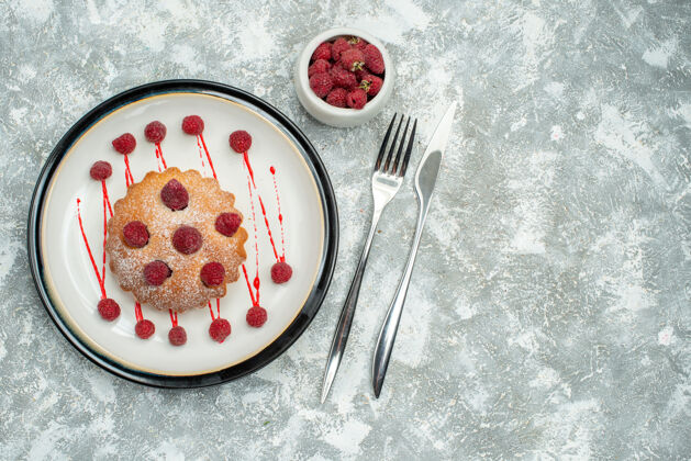 盘子顶视图浆果蛋糕在白色椭圆形盘子上 覆盆子在碗叉和餐刀在灰色表面自由空间椭圆形粉末浆果蛋糕