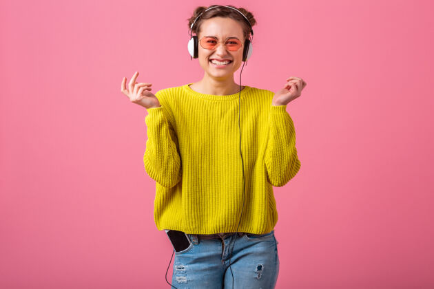 时尚迷人的快乐有趣的女人戴着耳机听音乐穿着时髦的五颜六色的服装隔离在粉红色的墙上 穿着黄色的毛衣和太阳镜 玩得很开心有趣微笑休闲