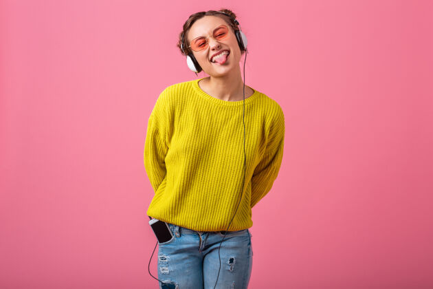 耳机迷人的快乐有趣的女人戴着耳机听音乐穿着时髦的五颜六色的服装隔离在粉红色的墙上 穿着黄色的毛衣和太阳镜 玩得很开心夏天漂亮快乐