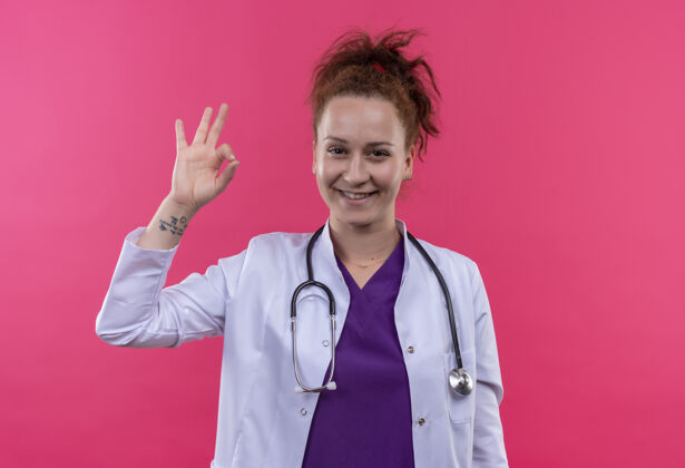 站着年轻的女医生穿着白大褂 手持听诊器 面带微笑 脸上挂着“一切正常”的牌子 站在粉红色的墙上微笑穿上脸